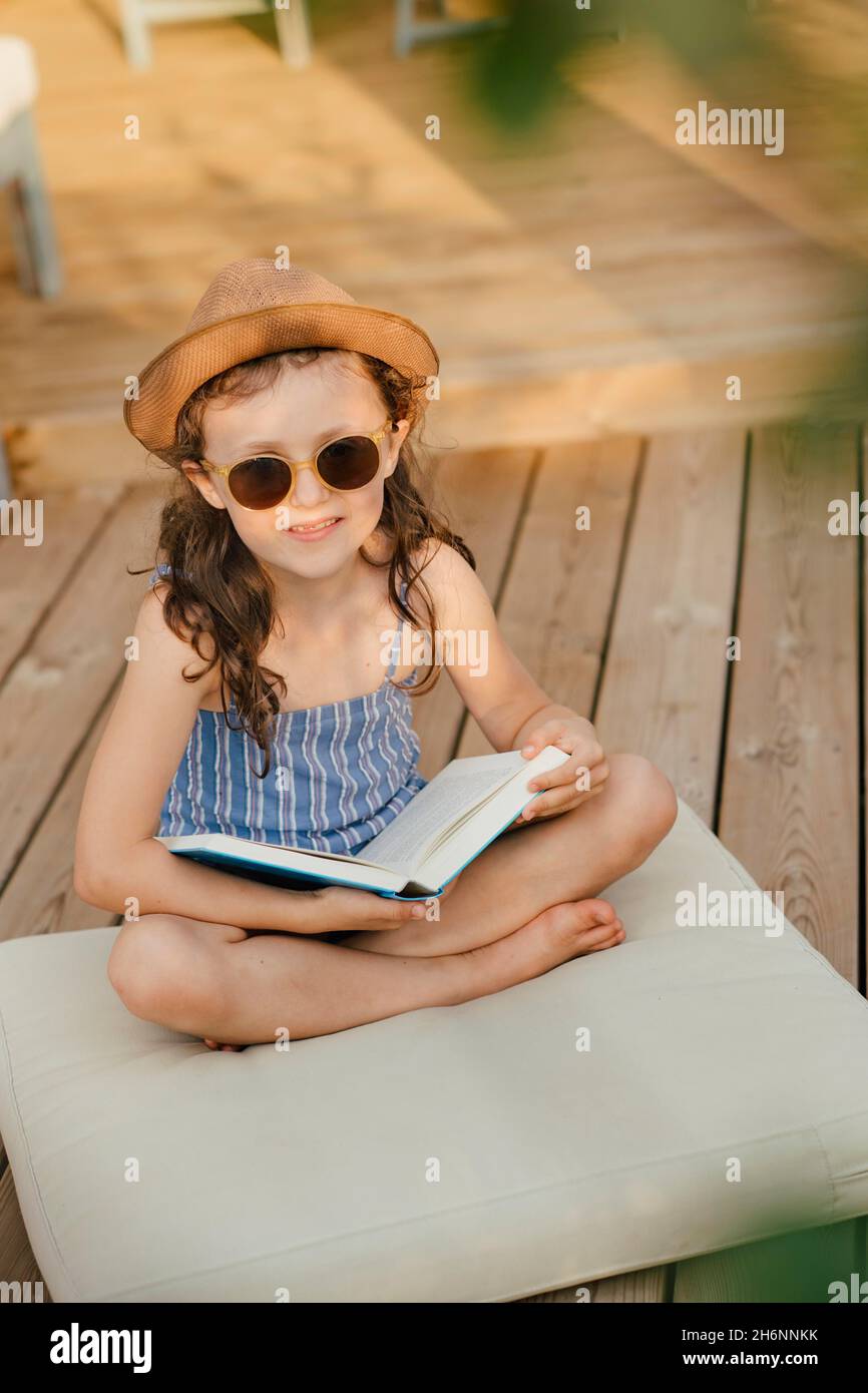 Mädchen mit Sonnenbrille sitzt auf dem Kissen auf der Holzterrasse und liest ein Buch Stockfoto