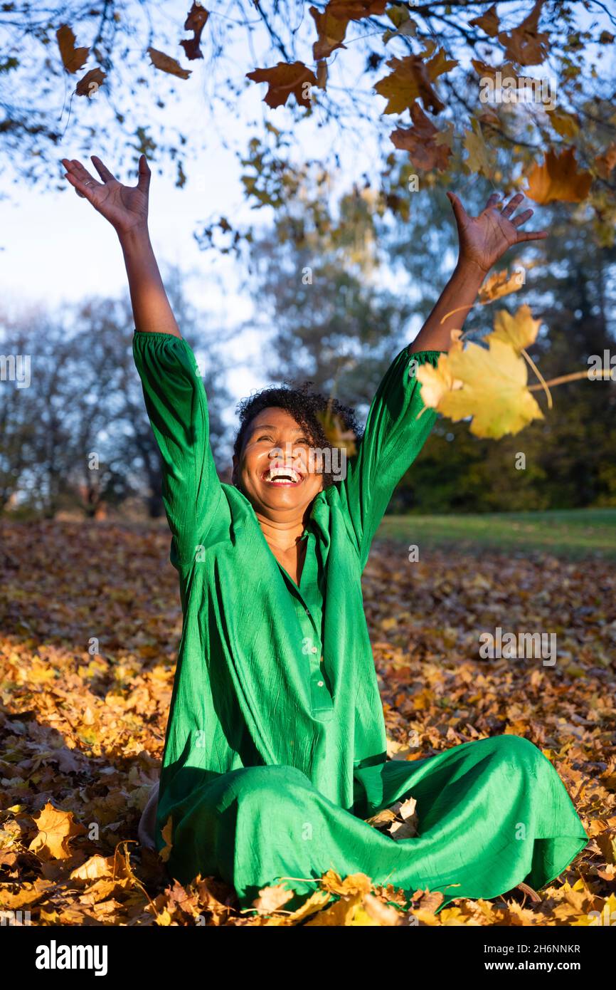 Porträt einer lachenden, dunkelhäutigen Frau mit Locken in einem grünen Kleid, sitzend und Herbstblätter werfend, Außenaufnahme im Herbst, Deutschland Stockfoto