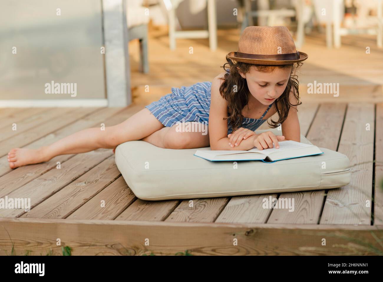 Mädchen, das auf dem Kissen auf der Holzterrasse sitzt und ein Buch liest Stockfoto