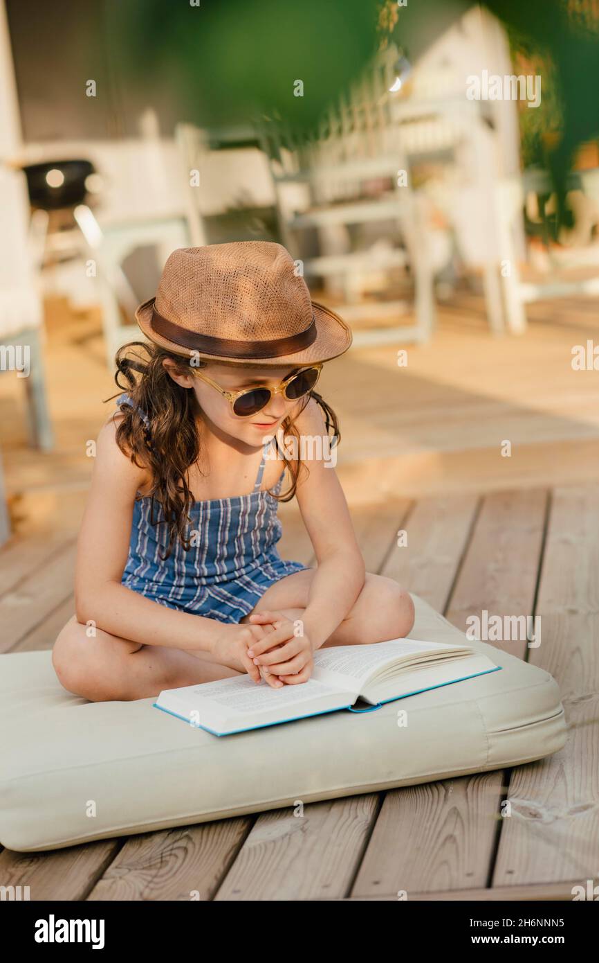 Mädchen mit Sonnenbrille sitzt auf dem Kissen auf der Holzterrasse und liest ein Buch Stockfoto