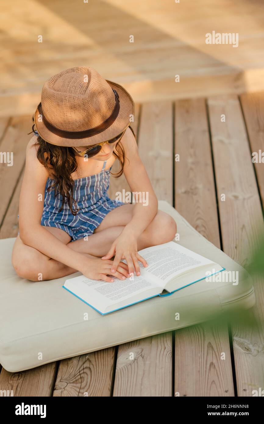 Mädchen mit Sonnenbrille sitzt auf dem Kissen auf der Holzterrasse und liest ein Buch Stockfoto