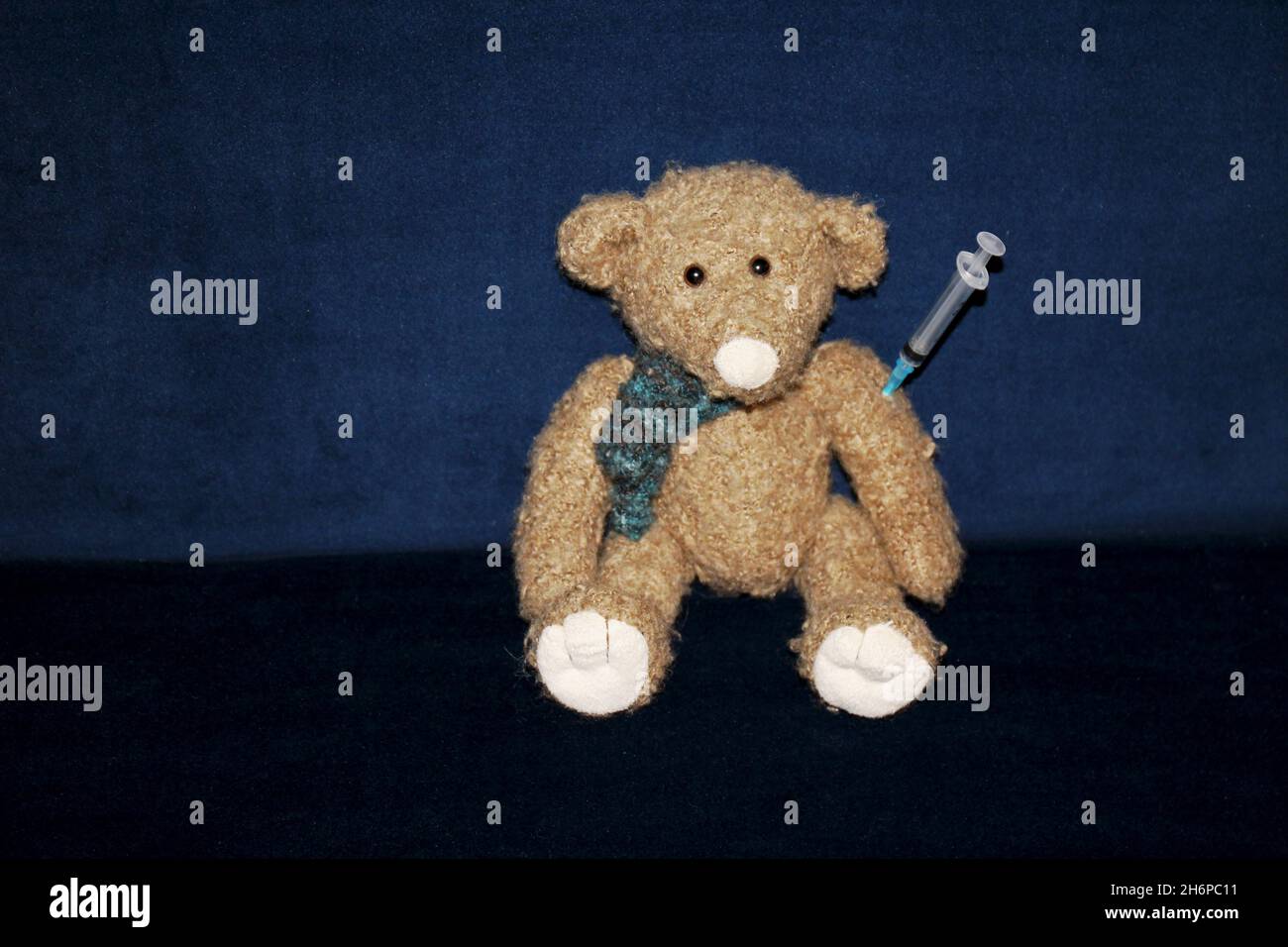 Kuscheltier Teddy mit Spritze im Arm, sitzend auf einem dunkelblau Sofa aus Samt. Stockfoto