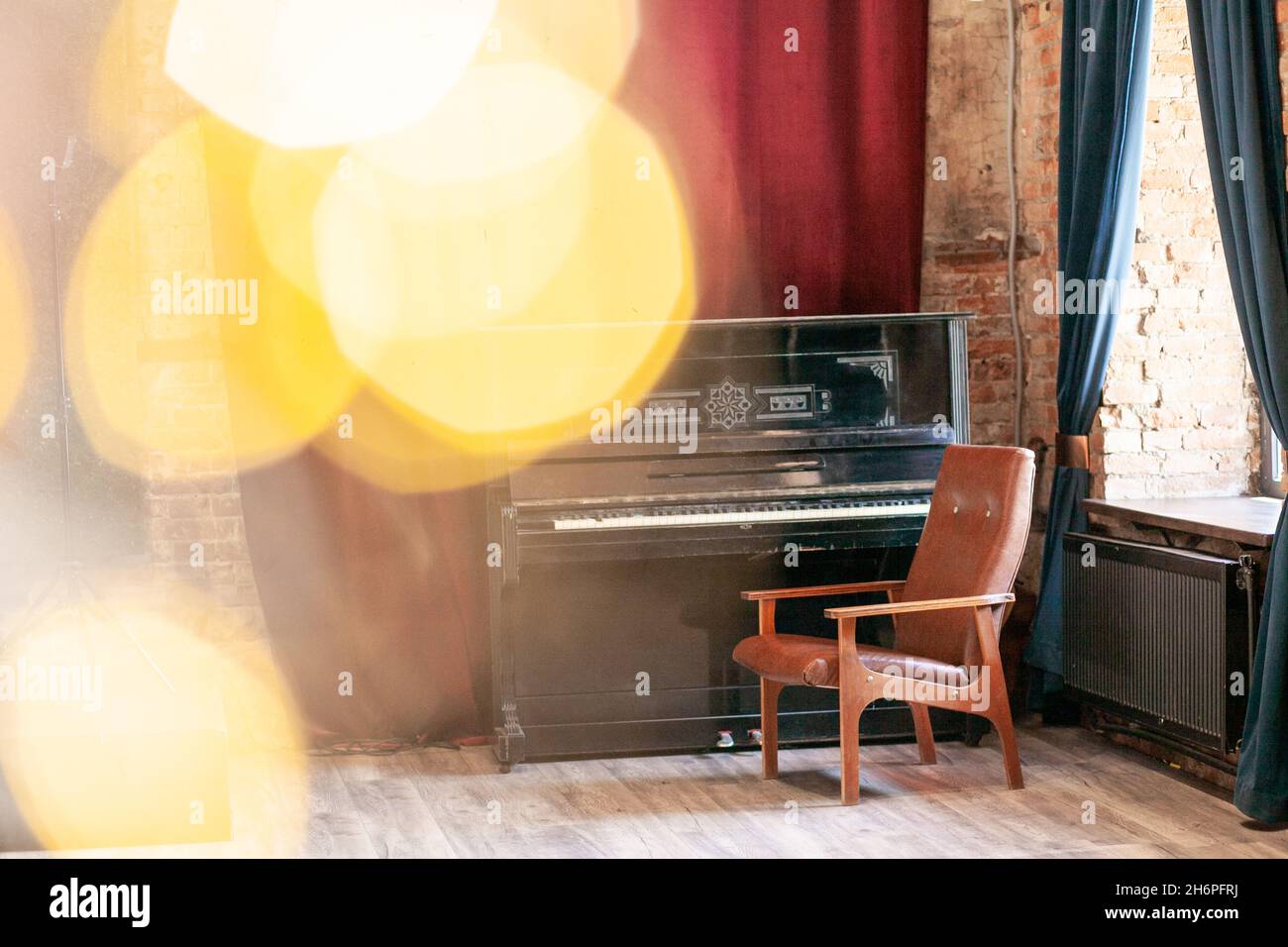 Altes Vintage-Klavier und Sessel stehen in der Halle vor dem Hintergrund eines roten Vorhangs und einer Backsteinmauer in der Nähe des großen Fensters, mit Bokeh und Highlig Stockfoto