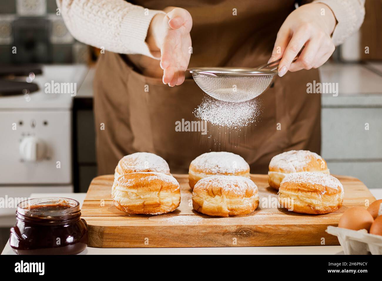 Frau bereitet frische Donuts mit Marmelade in der heimischen Küche. Kochen traditionelle jüdische Hanukkah sufganiyot. Hände bestreuen Berliner mit Puderzucker. Stockfoto