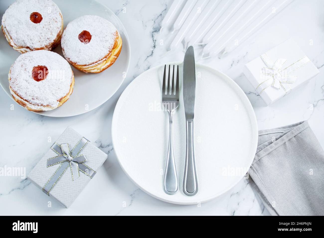 Tischeinstellung für den jüdischen Feiertag der Chanukka. Teller und Besteck auf weißem Hintergrund. Kerzen, Geschenke und Dessert Donut sufganiyot. Stockfoto