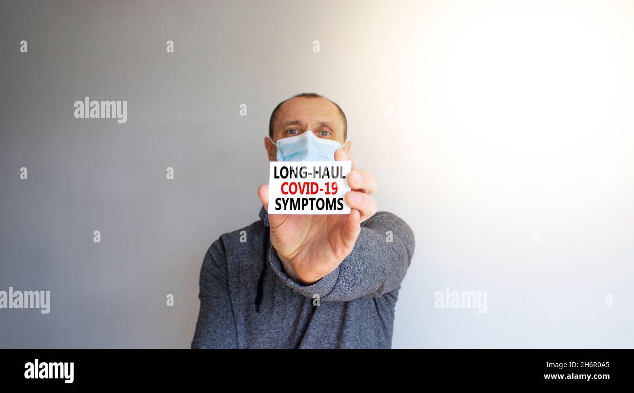 Symbol für COVID-Symptome bei Langstrecken-Kovidsymptome. Weiße Karte, Worte Langstrecke covid-19 Symptome. Ein junger Mann in grauer Kleidung und medizinischer Maske. Schöner weißer Hintergrund Stockfoto