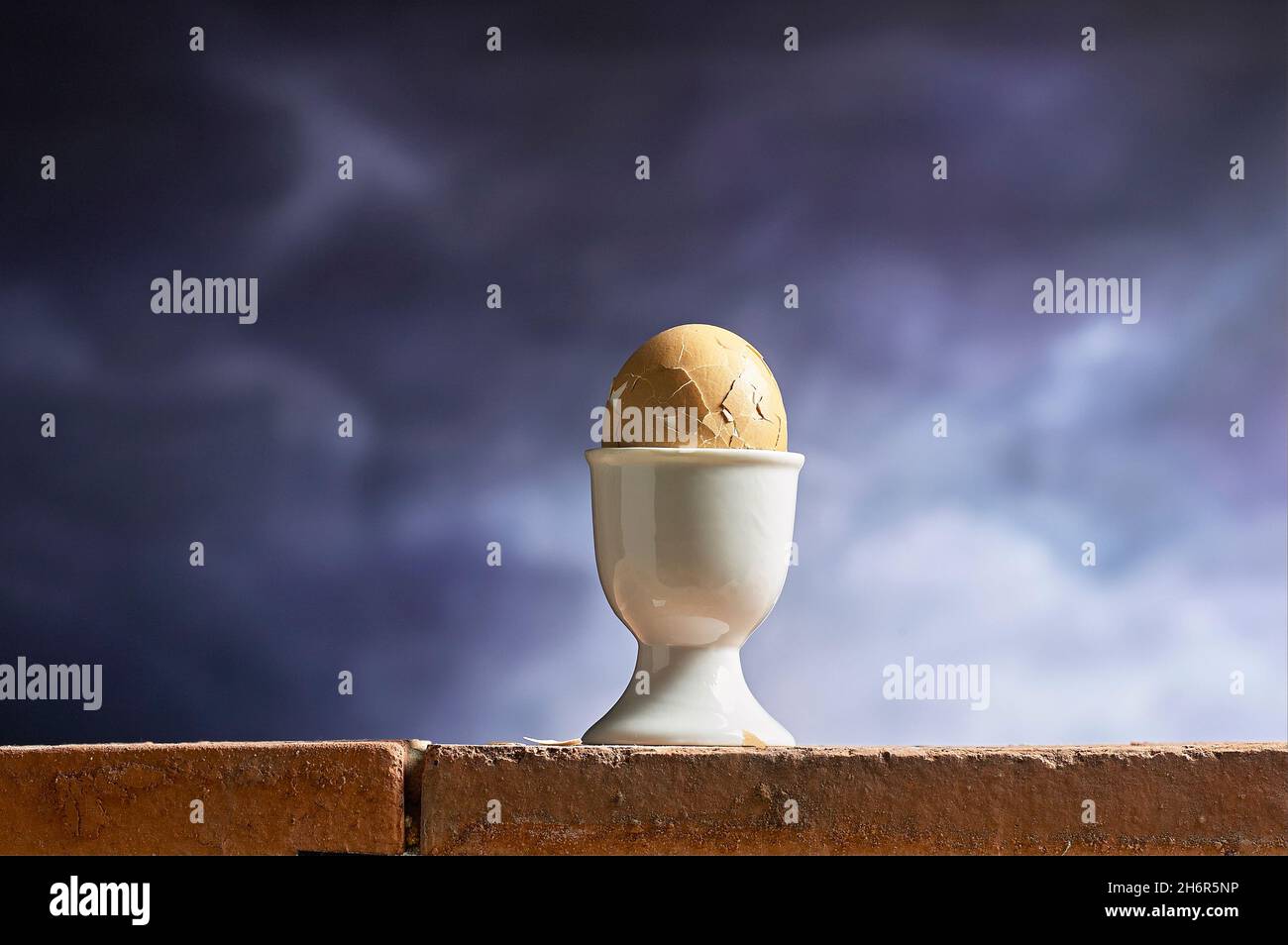 Zertrümmertes gekochtes Ei in einem Eierbecher-Halter, der wie humpty dumpty auf der Ziegelwand positioniert ist. Stockfoto