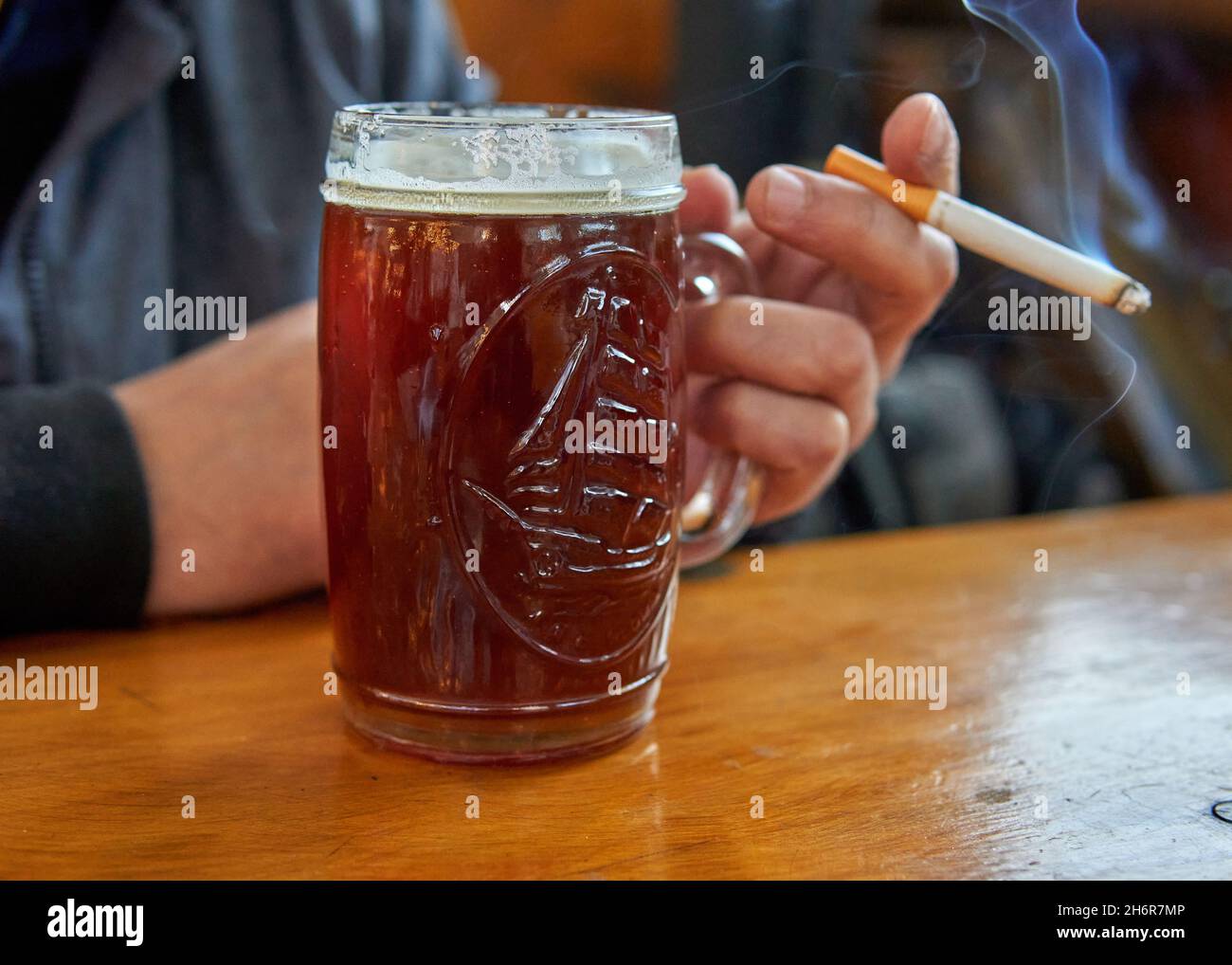 Die Hand eines Mannes hält einen Becher Bier und eine Zigarette auf einem Holztisch. Genießen Sie das Rauchen und Alkohol trinken. Horizontal. Selektiver Fokus Stockfoto