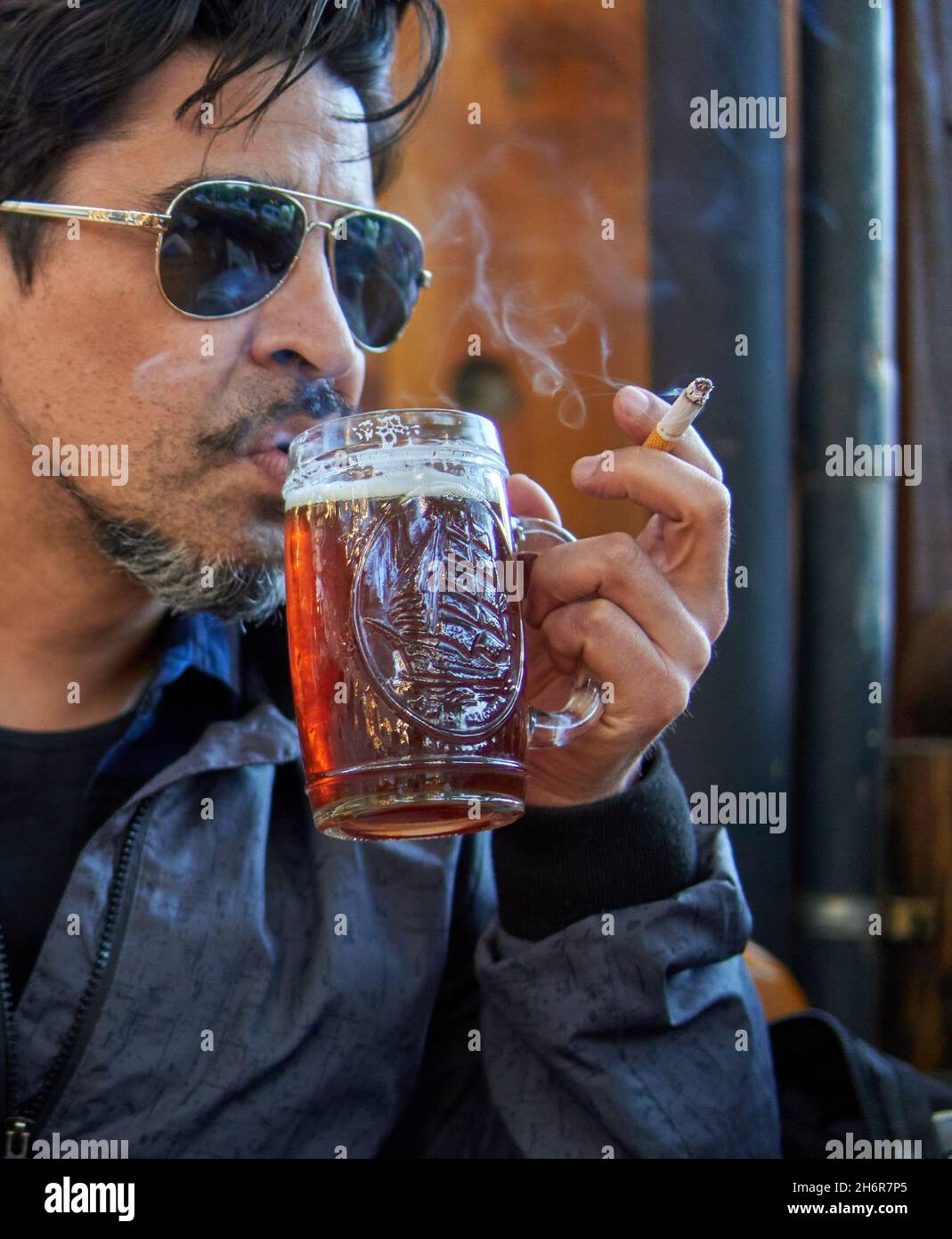 Brunette latin Mann im Profil mit Sonnenbrille trinken ein Pint Amber Craft Bier und rauchen eine Zigarette im Urlaub in einer Bar. Vertikal Stockfoto