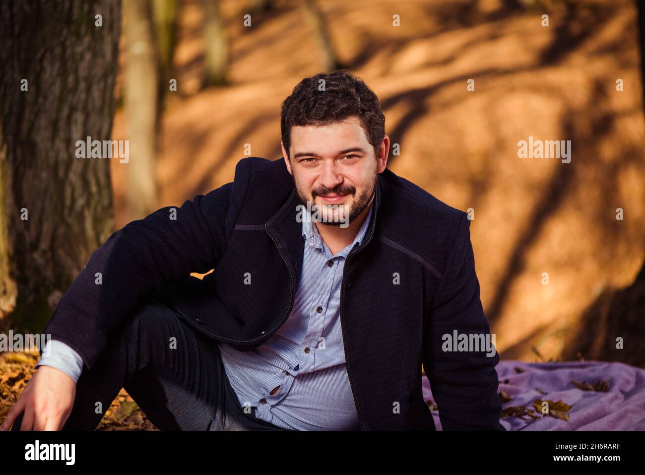 Junger Mann im Mantel Porträt im Herbstwald mit Bäumen Schatten auf dem Hintergrund Stockfoto