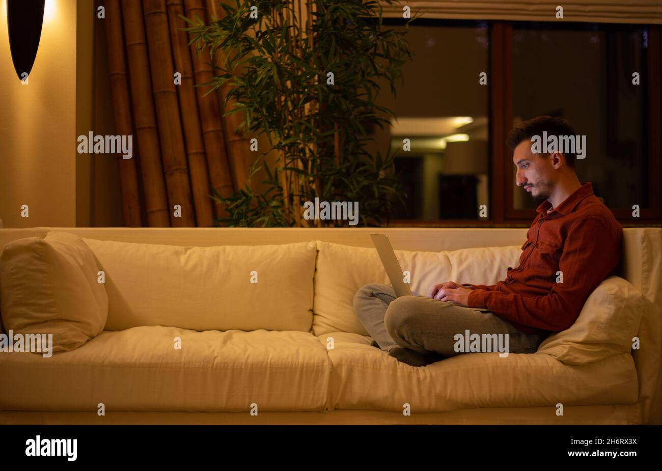 Ein junger Mann mit einem Bart in einem roten Hemd arbeitet mit einem Laptop und sitzt abends im Haus auf der Couch. Stockfoto