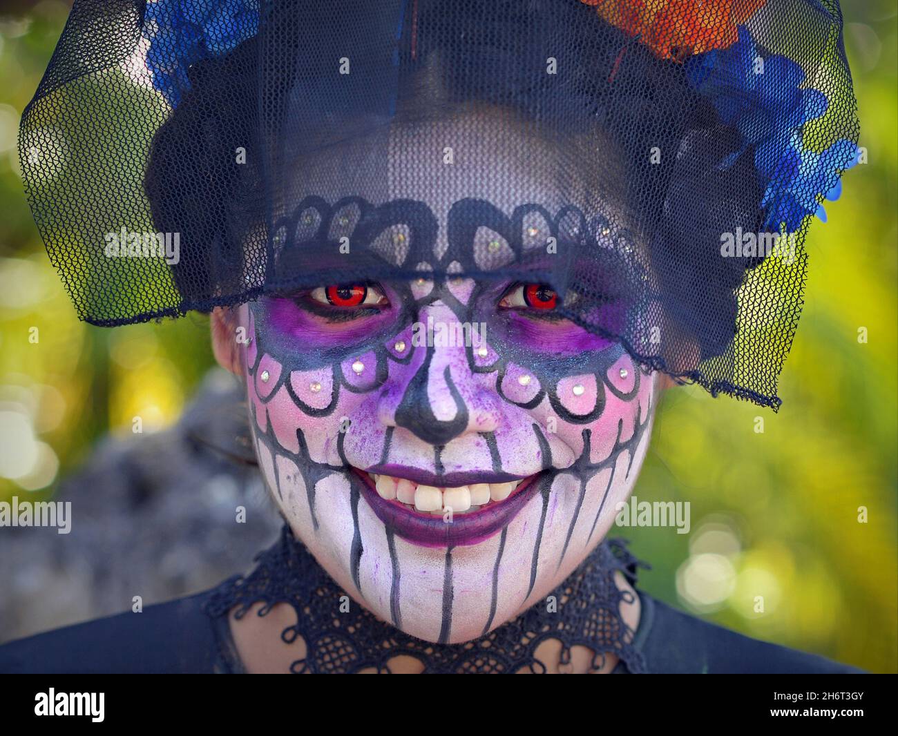 Kostümierte junge, hübsche mexikanische Yucatecan-Frau mit Gesichtsbemalung und orangefarbenen Kontakten lächelt den Betrachter am Tag der Toten (Día de los Muertos) an. Stockfoto