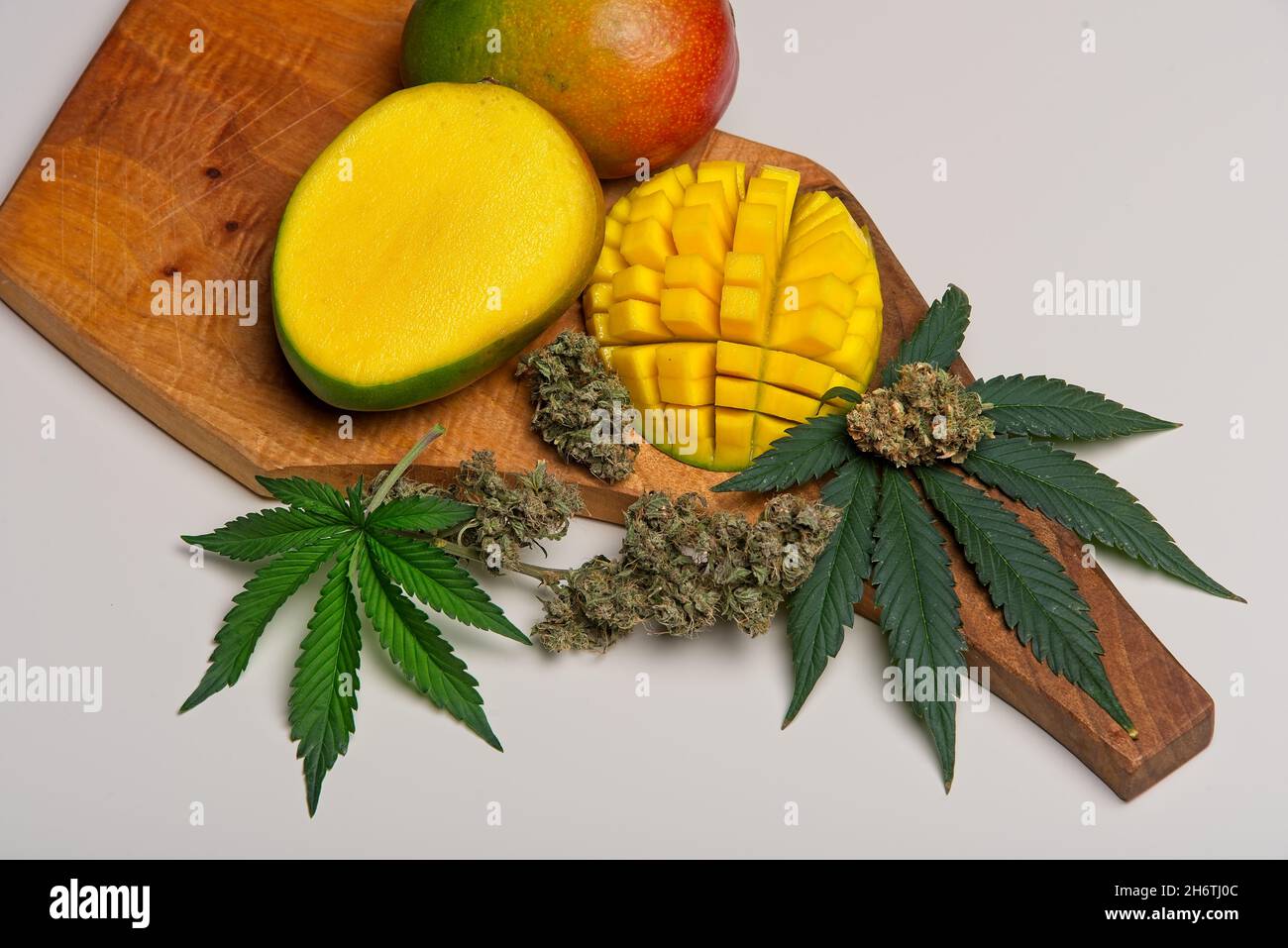 Draufsicht auf geschnittene Mangofrüchte mit Cannabisblättern und Knospen. Myrcen und Terpene in Mangos unterstützen die Wirkungen von THC in Cannabis. Auf Holzschneidplatte Stockfoto