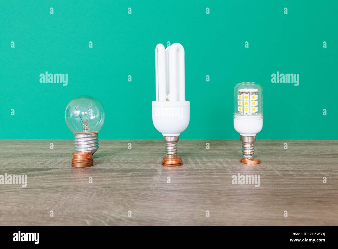 Drei Glühlampen, ein Wolframfaden, eine Energiesparlampe und eine LED-Glühlampe. Vor ihnen liegen Stapel von Münzen nach dem Preis ihrer Stockfoto