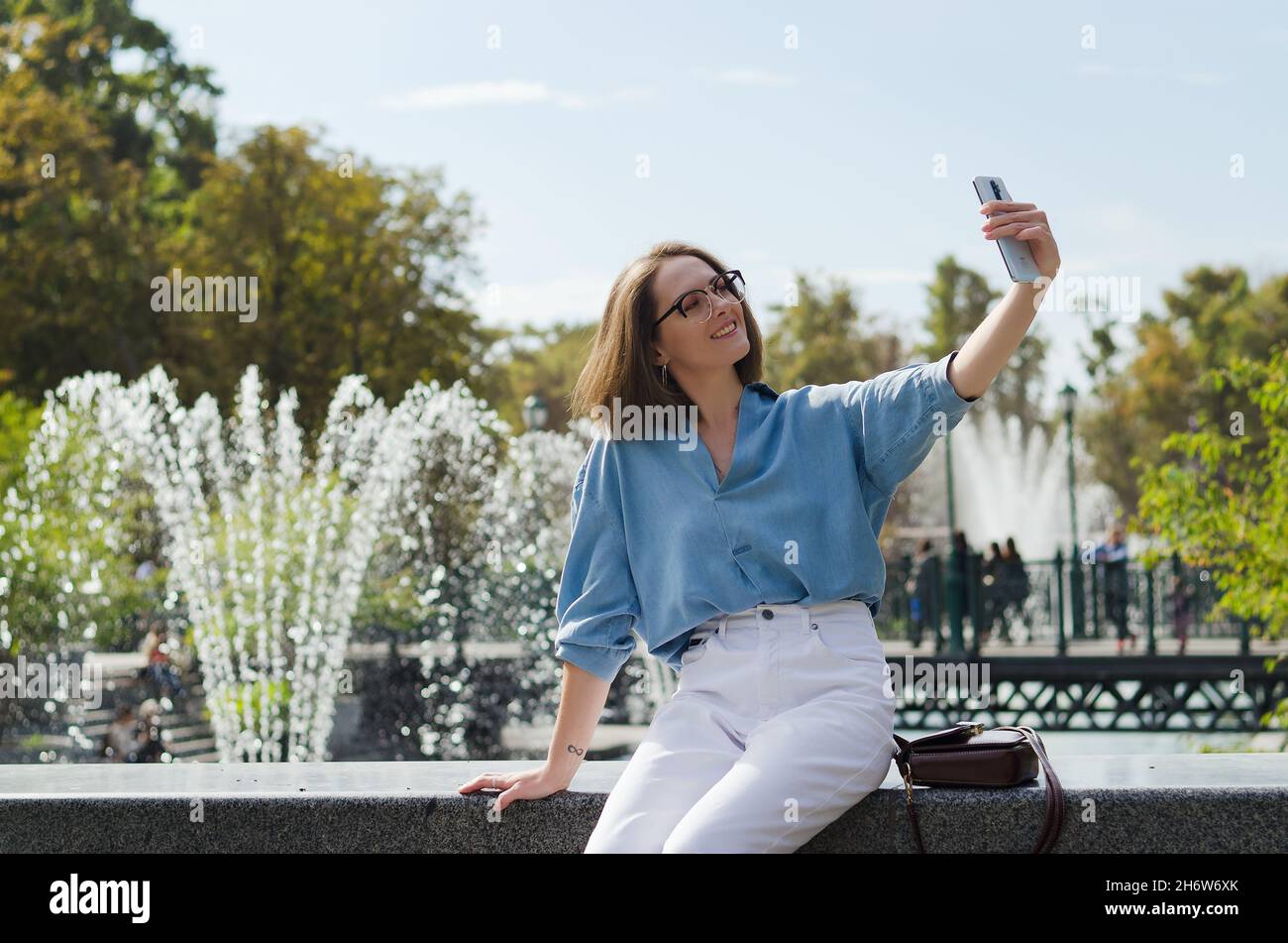Urbanes Porträt einer jungen attraktiven Geschäftsfrau in Brillen und legerer Kleidung. Frau, die in der Stadt läuft, im Park, telefoniert, Selfie macht Stockfoto