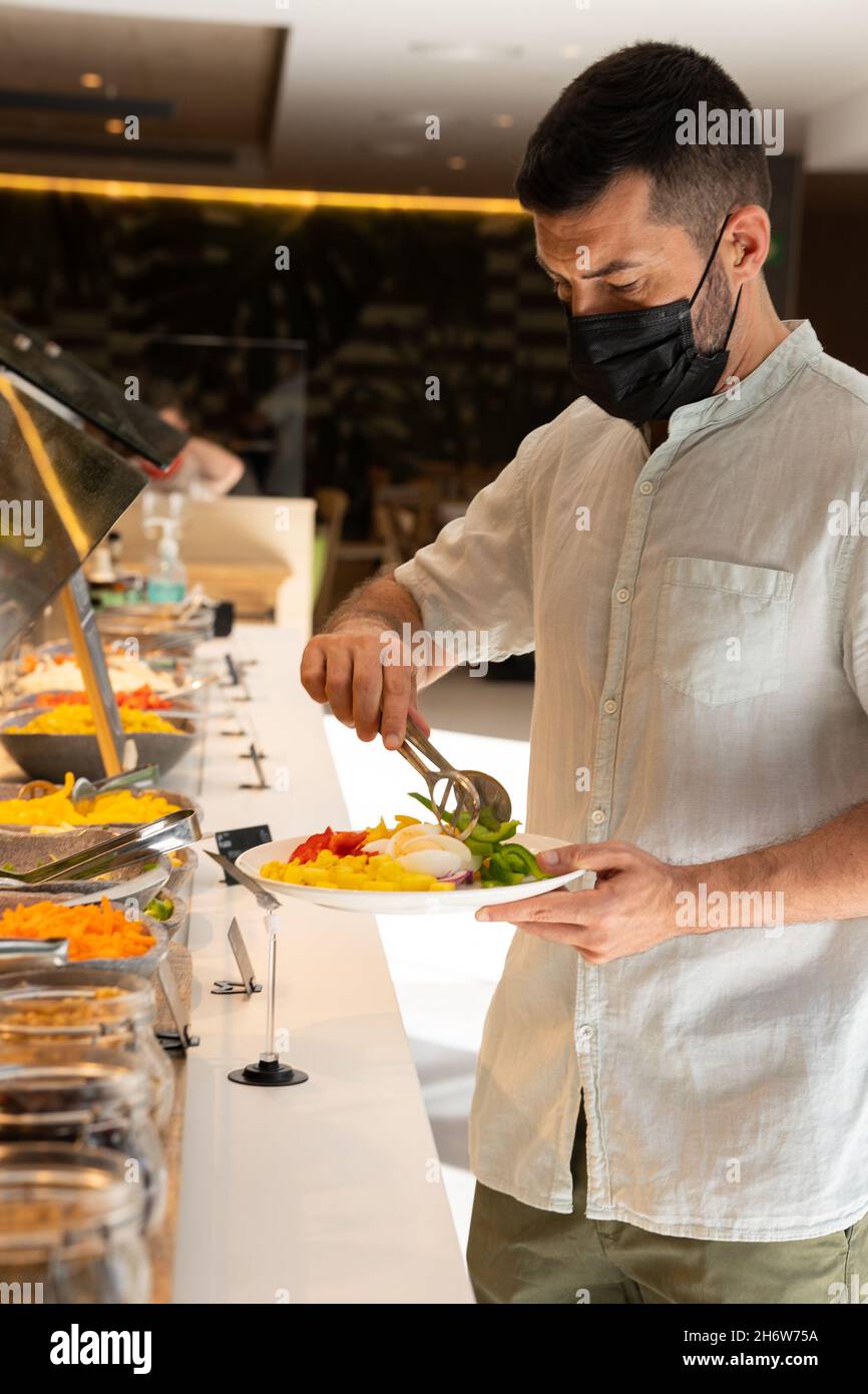 Mann mit Gesichtsmaske gießt sich selbst Essen in das Hotelbuffet. Stockfoto