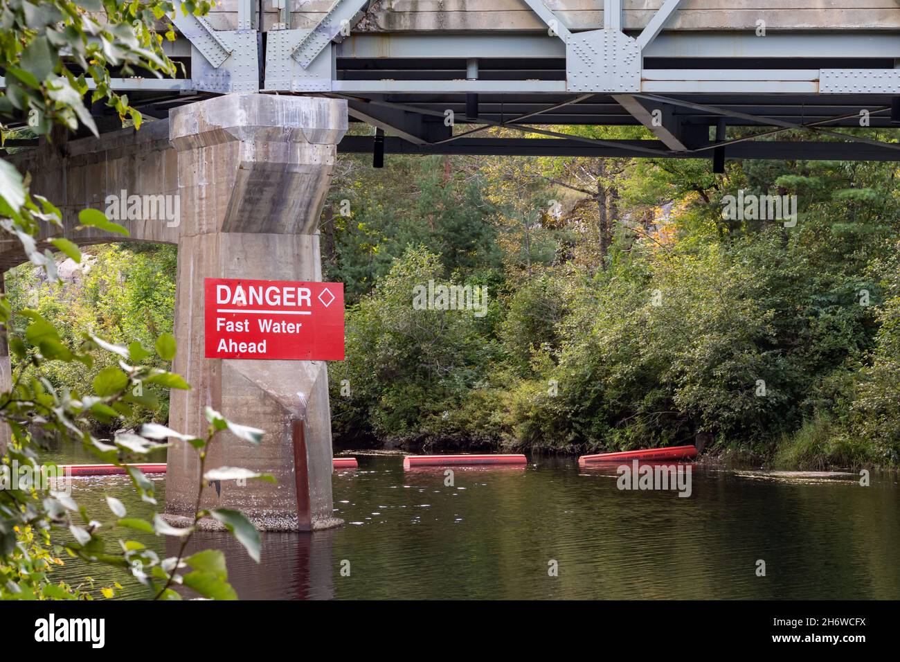 Rotes Warnschild am Brückenpfosten, das schnelles Wasser vor sich anzeigt. Rote Bojen im Fluss, um zu verhindern, dass Bootsfahrer in Stromschnellen eindringen. Stockfoto