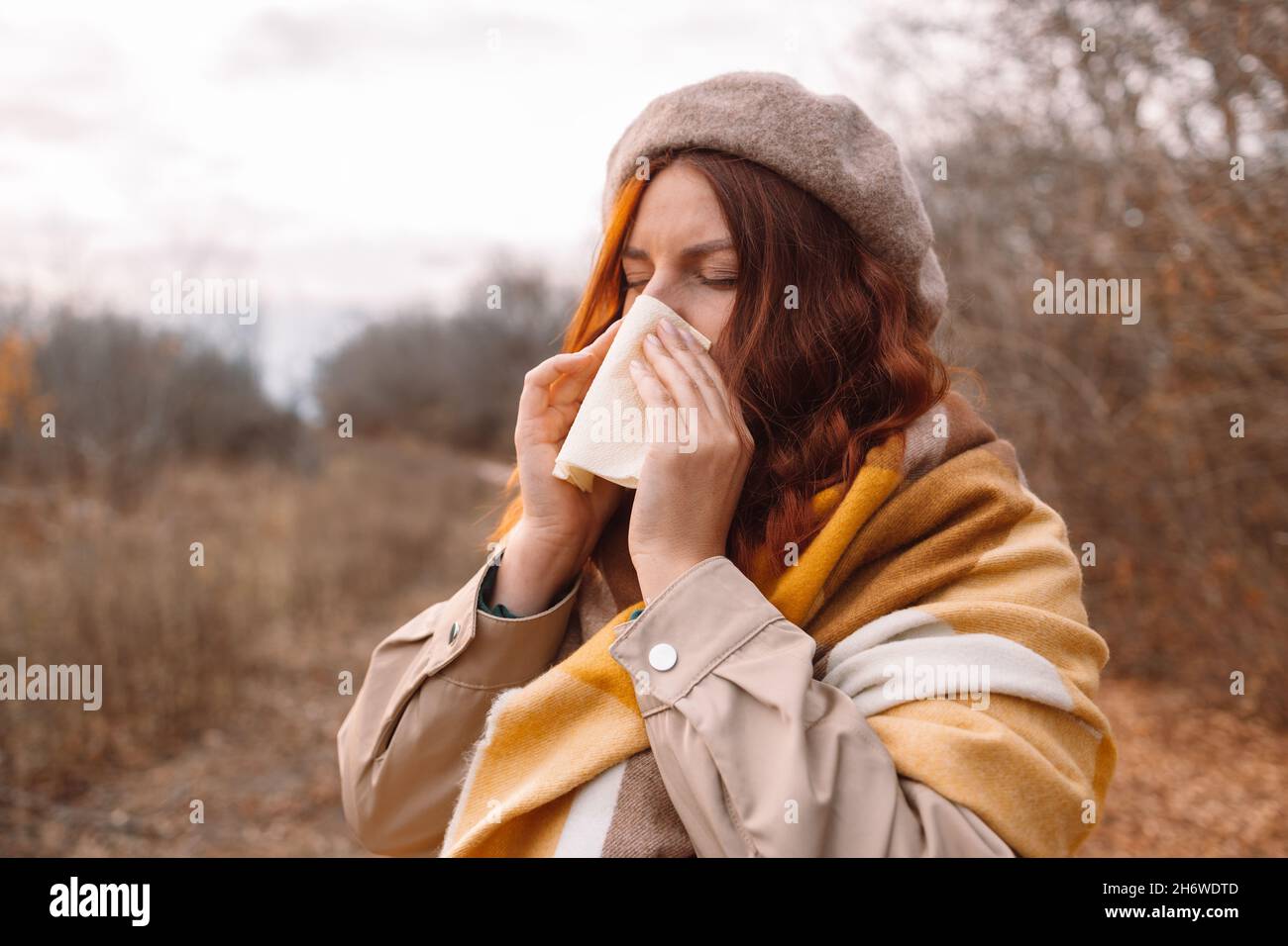 Kranke junge kaukasische Frau in stilvoller Kleidung mit Erkältung und Grippe steht im Freien, niest, wischt Nase mit Papierserviette im städtischen Herbstpark Stockfoto