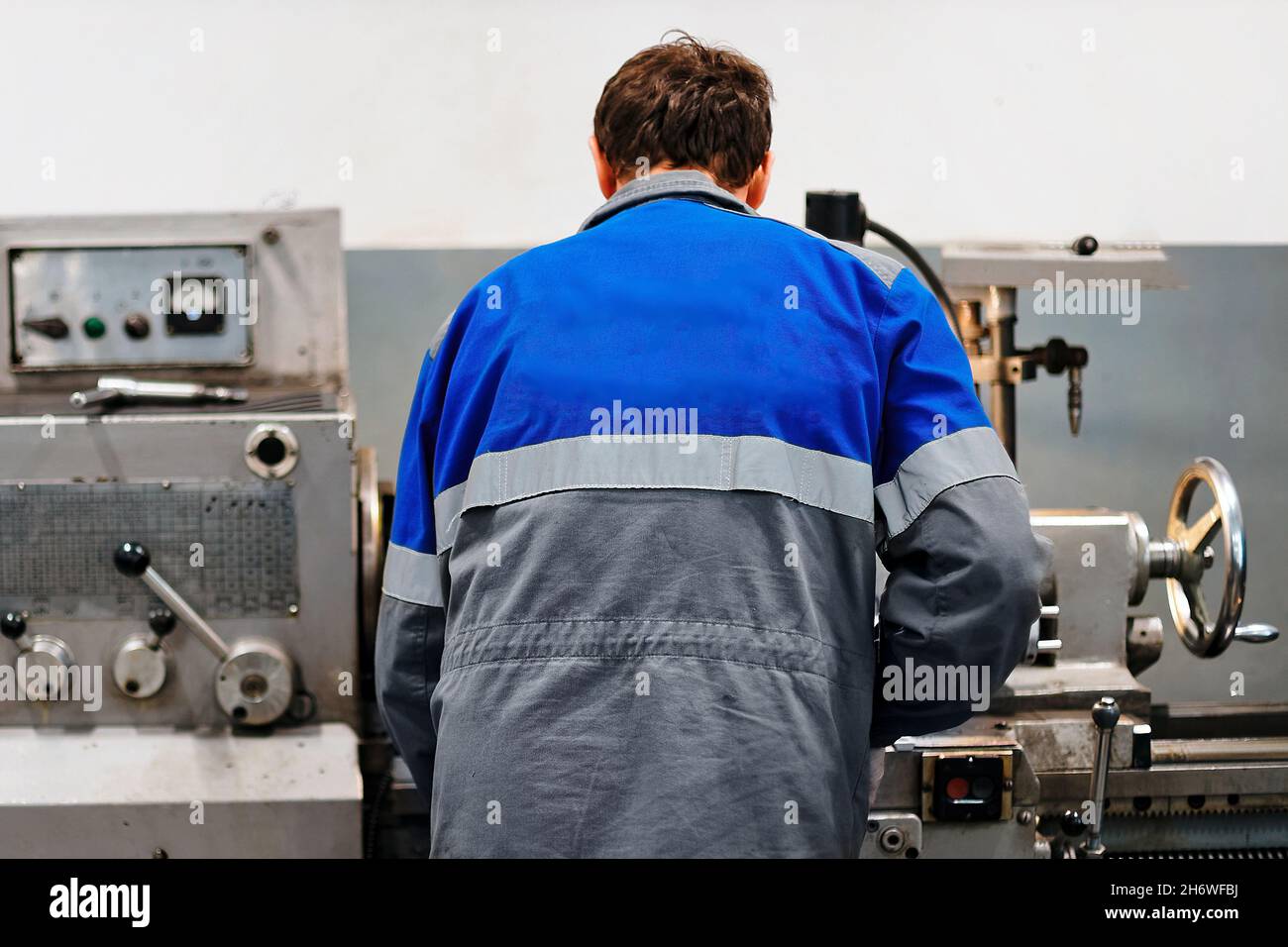 Turner steht hinter der Drehmaschine in der Produktionshalle und arbeitet. Blick auf den Arbeiter von hinten in Overalls. Authentische Arbeitsprozess-Szene in der Produktion. Echter Arbeiter. Nicht inszenierte Szene. Stockfoto