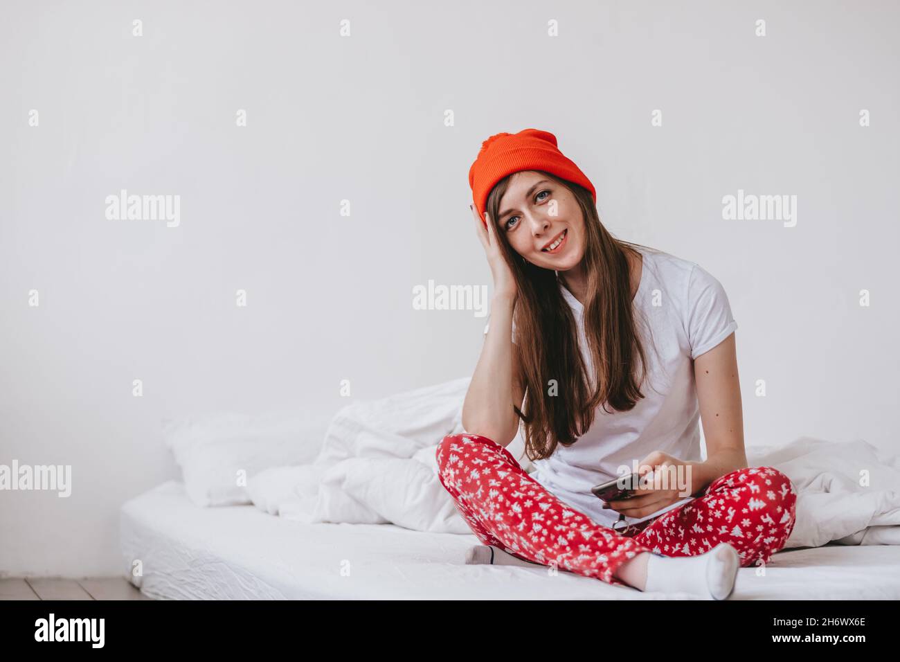 Lächelndes lustiges Mädchen in einem roten Hut und Weihnachten Pyjamas sitzen im Bett mit weißen Laken. Feiertage zu weihnachten. Gute Weihnachtsstimmung Mädchen. Stockfoto