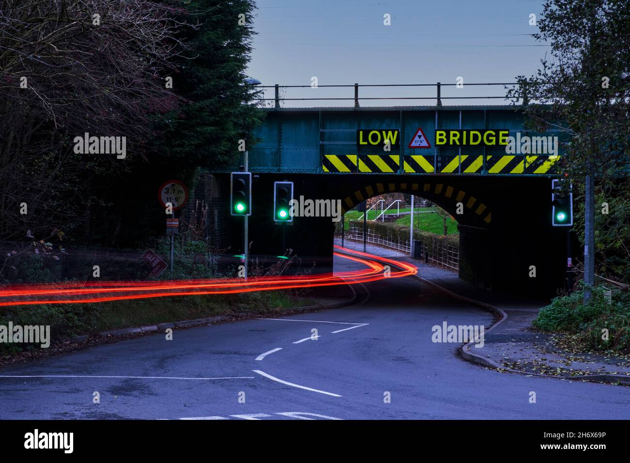 Auto mit Rotlichtweg unter Eisenbahnbrücke mit niedrigem Brückenwarnschild in der Dämmerung Stockfoto