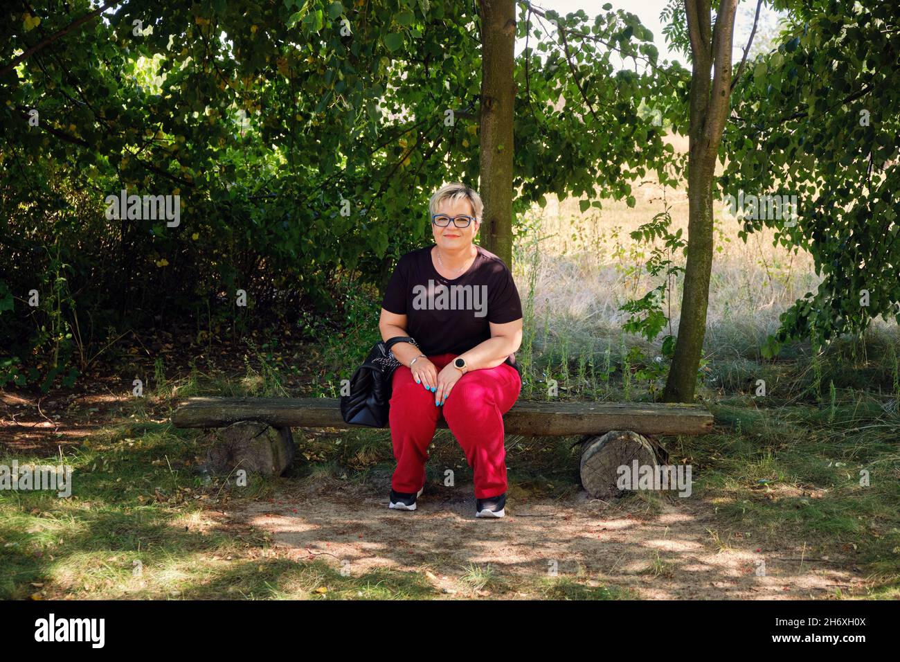 Ganztägige Ansicht einer reifen Frau, die an einem sonnigen Tag in Polen auf einer hölzernen Parkbank sitzt Stockfoto