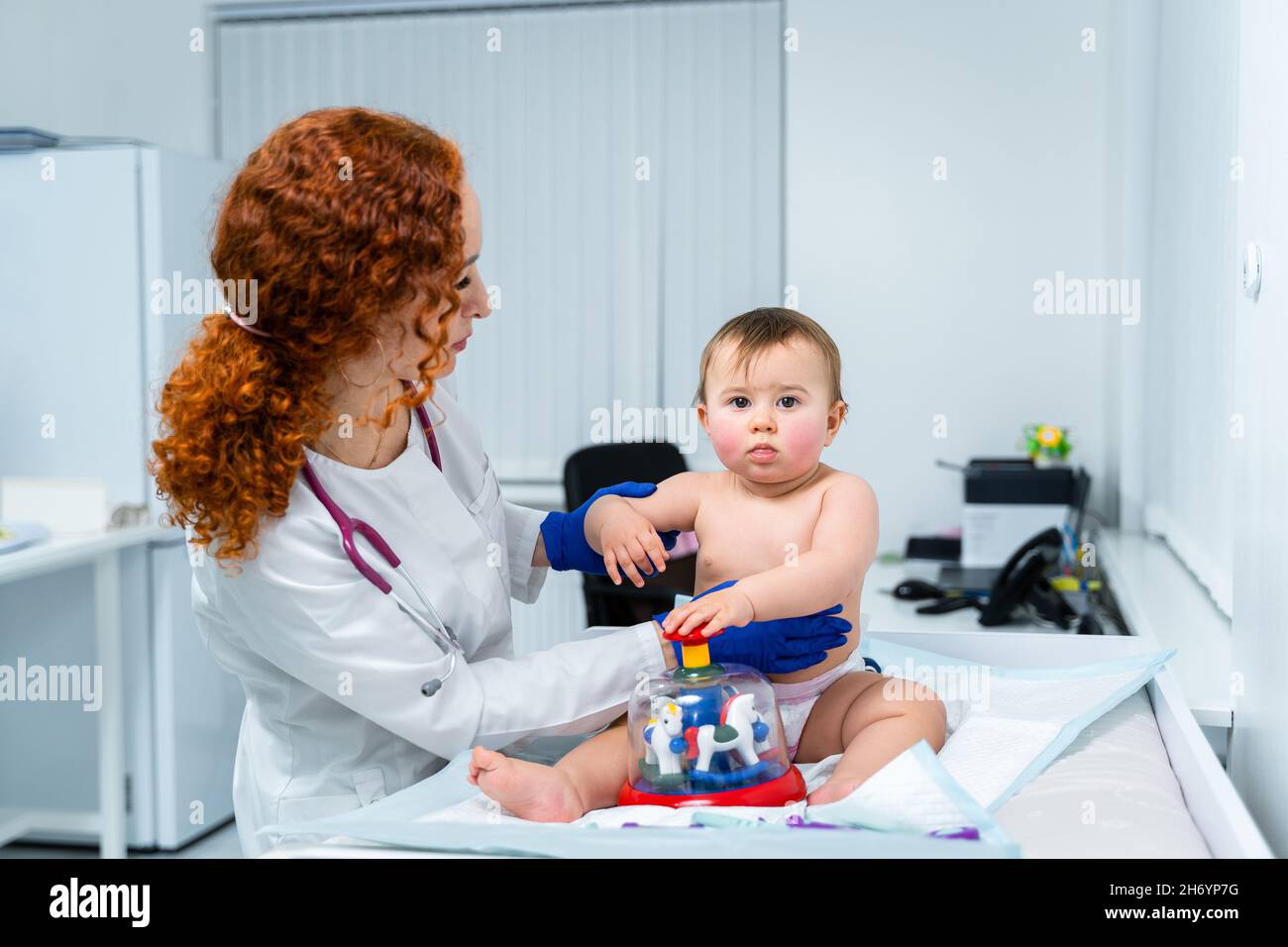 Eine rothaarige Ärztin untersucht das kleine Kind weniger als ein Jahr lang in einer modernen Klinik. Kinderarzt während der Untersuchung Baby im Krankenhaus Stockfoto