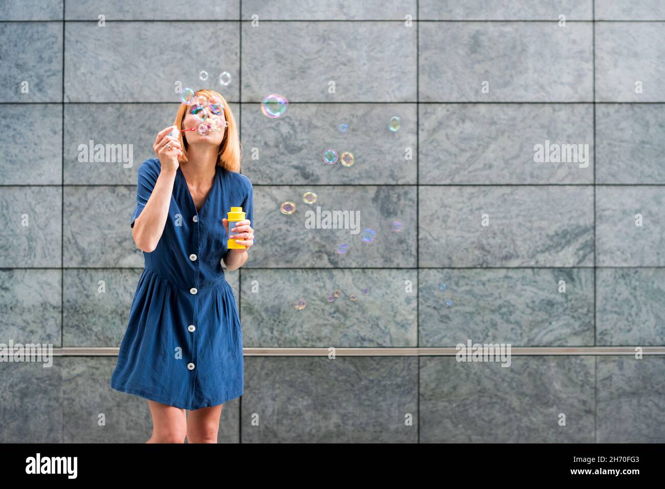 Sorglose junge Frau in blauem Kleid weht Regenbogen Seifenblasen während Spaß am Sommertag vor grauen Wand Hintergrund Stockfoto