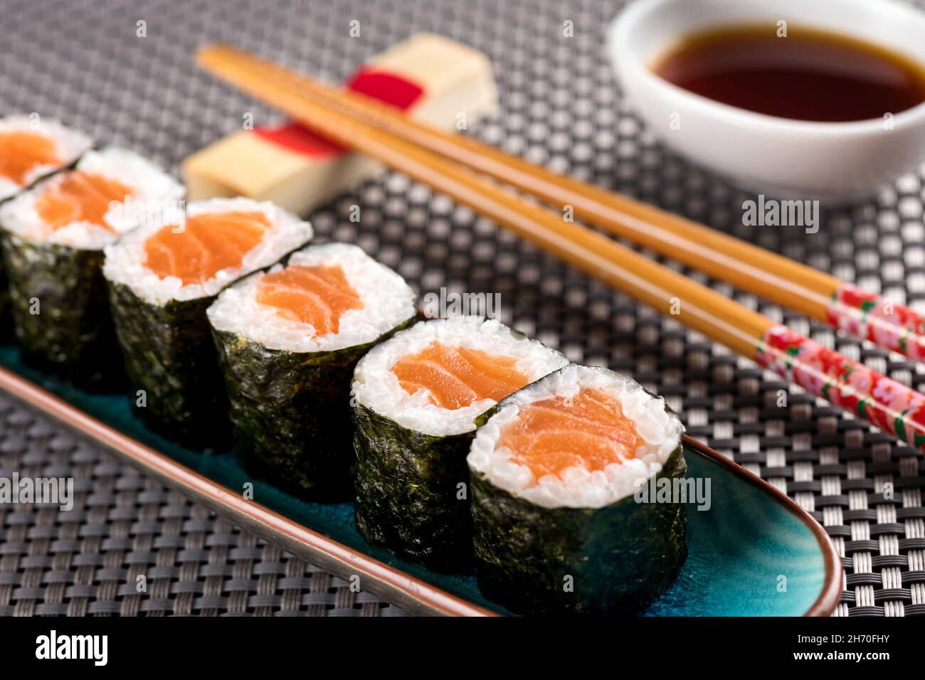 Lachs-Hosomaki-Sushi, serviert im Restaurant, mit Essstäbchen und Sojasauce, aus nächster Nähe auf einer gefroteten Bambusmatte Stockfoto