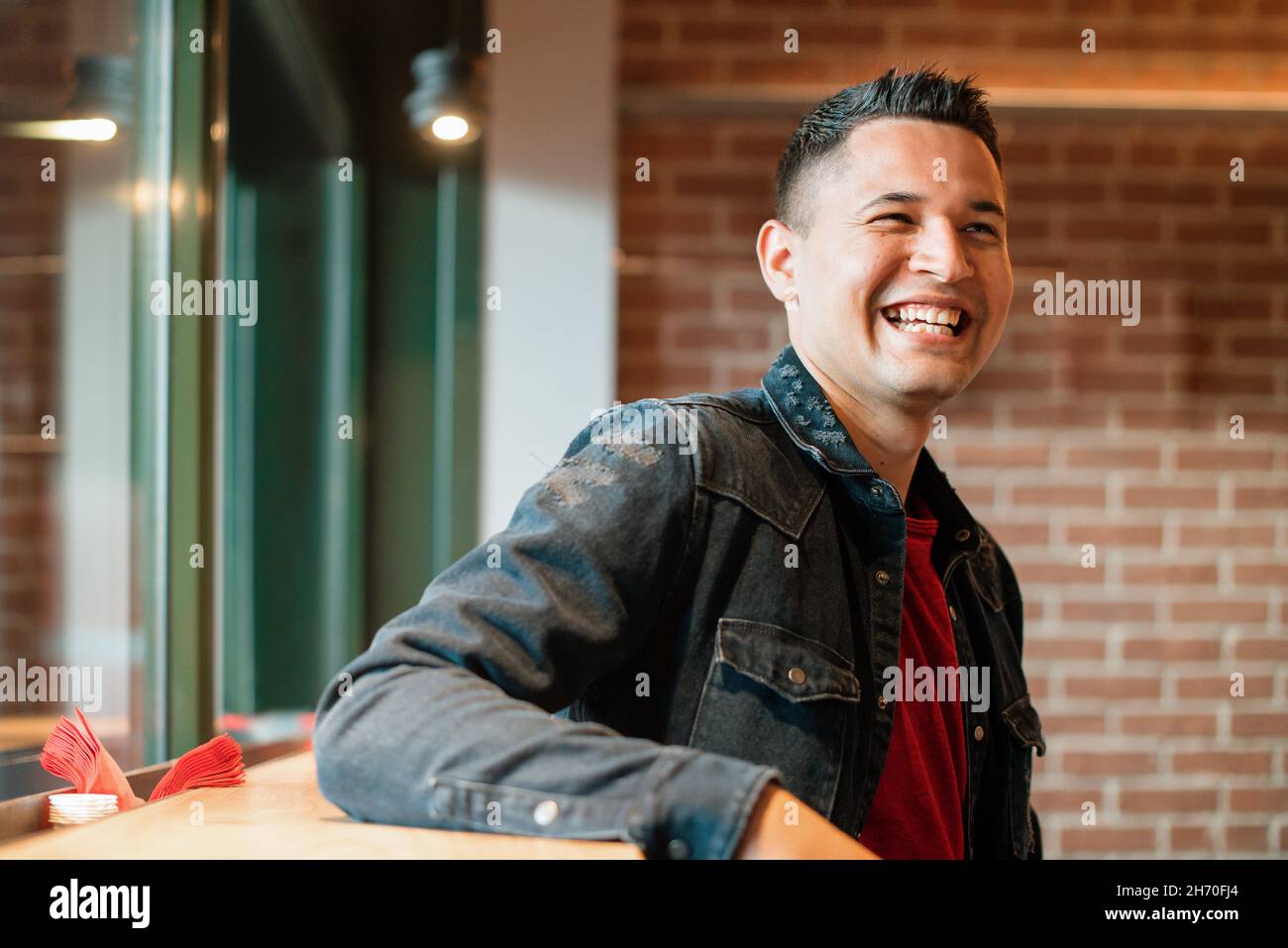 Fröhlicher junger hispanischer Kerl in Denim-Jacke, der hell lächelt, während er sich in einem Café im Loft-Stil mit Backsteinmauer in der Nähe des Fensters ausruhte Stockfoto