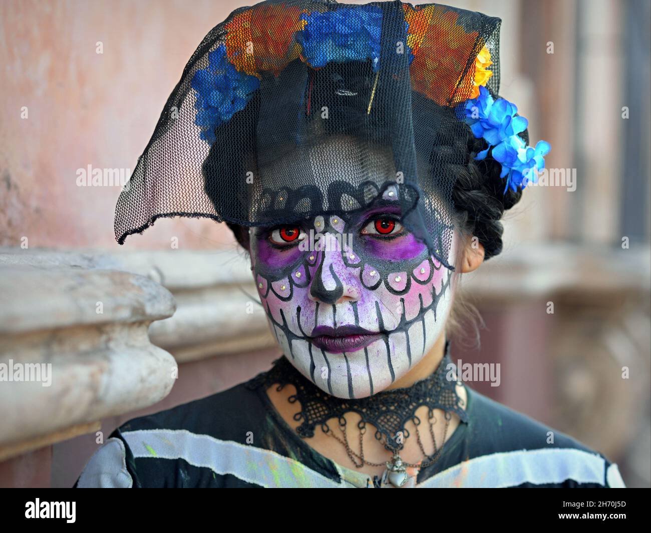 Kostümierte junge Mexikanerin mit gemaltem Gesicht und orangefarbenen Kontaktlinsen trägt am Tag der Toten (Día de los Muertos) einen schwarzen Vogelkäfig-Schleier. Stockfoto