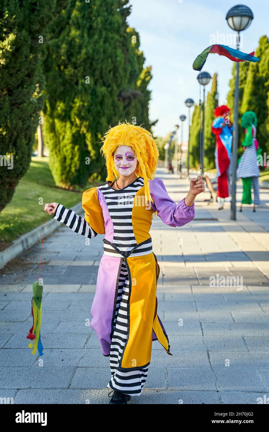 Fröhliche Clownin mit gemaltem Gesicht in lustigem Kostüm und gelber Perücke, lächelnd und auf dem Weg im Park mit bunten Poi in den Händen während der Parade Stockfoto
