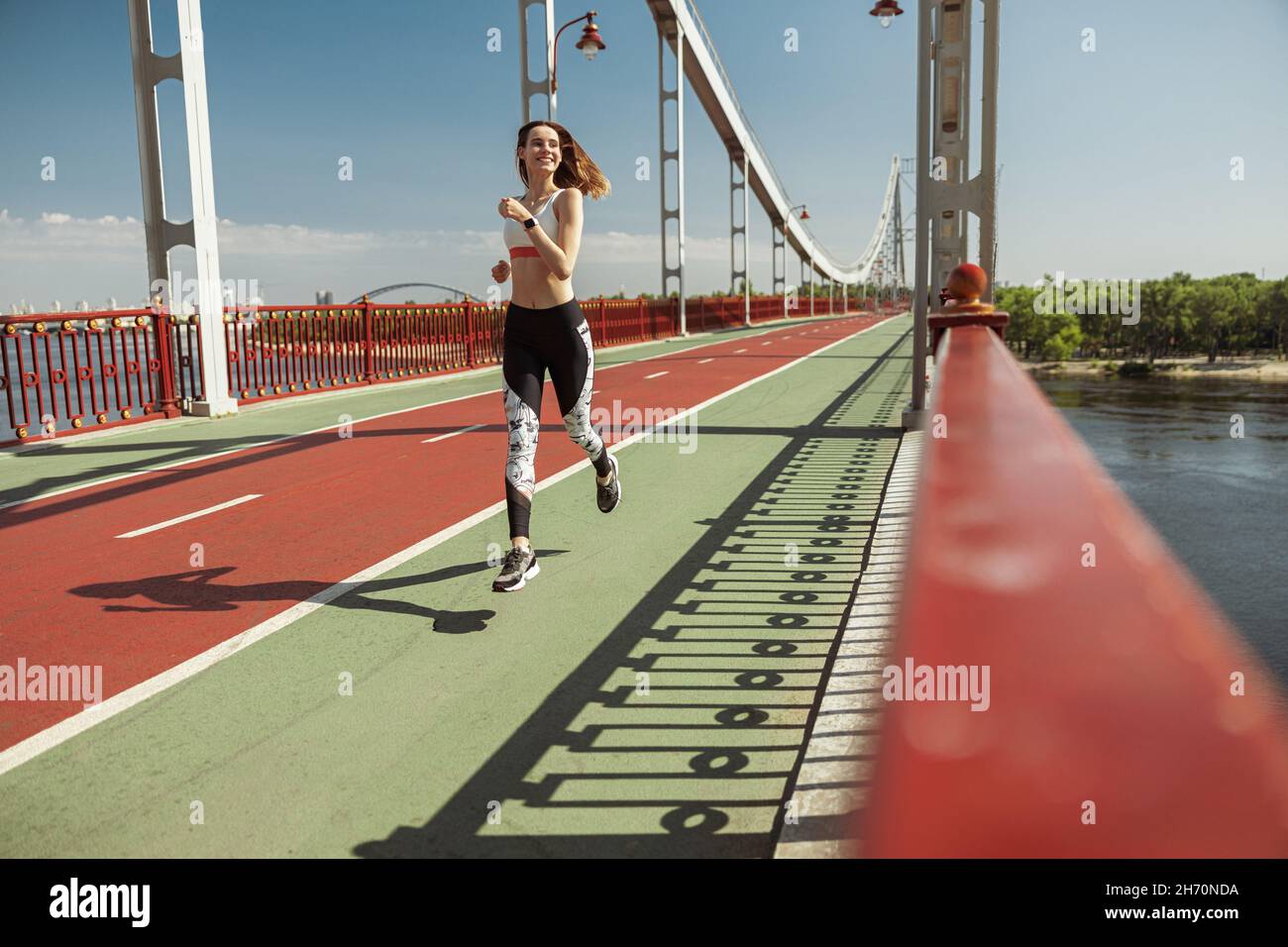 Lächelnde Dame mit lockeren Haaren läuft auf einem leeren Steg entlang der grünen Straße Stockfoto