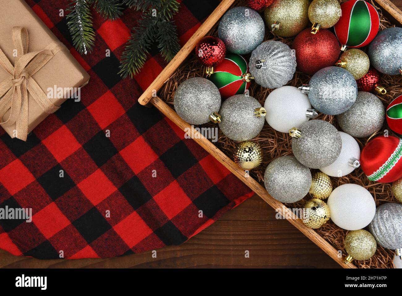 Christmas Flat Lay - Holzkiste mit Weihnachtsbaum-Ornamenten auf einem karierten Trre-Rock mit Geschenk und Kiefernästen. Stockfoto