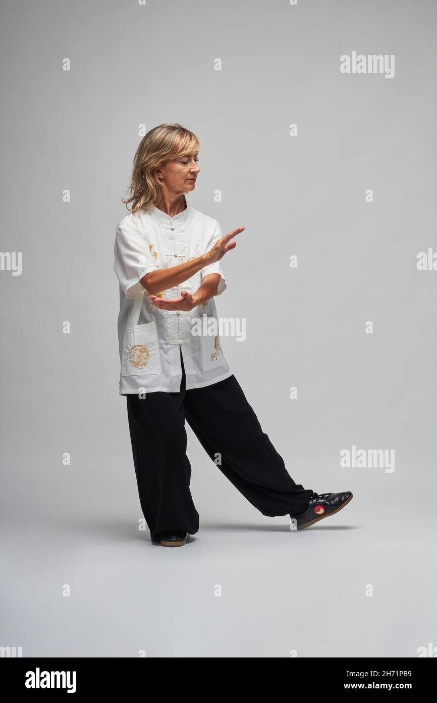 Reife blonde Frau, die Chi Kung und Tai Chi auf weißem Hintergrund praktiziert. Sie trägt eine traditionelle weiße chinesische Tai Chi Jacke, schwarze Hosen und BL Stockfoto