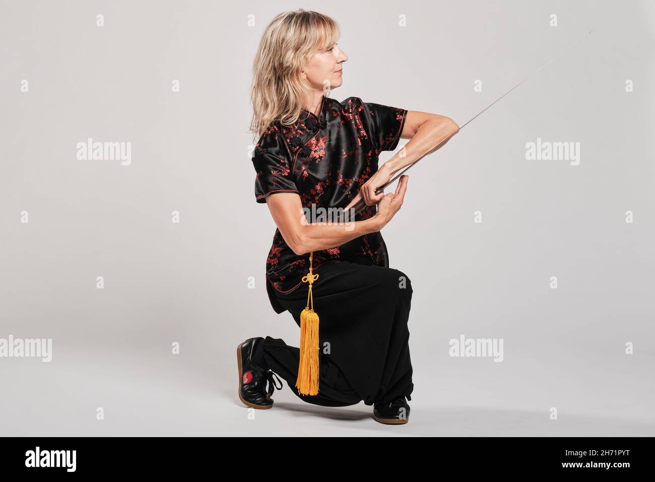 Reife blonde Frau, die Tai Chin mit Schwert auf weißem Hintergrund praktiziert, trägt eine traditionelle chinesische schwarze Jacke mit roter chinesischer Dekoration, schwarz Stockfoto