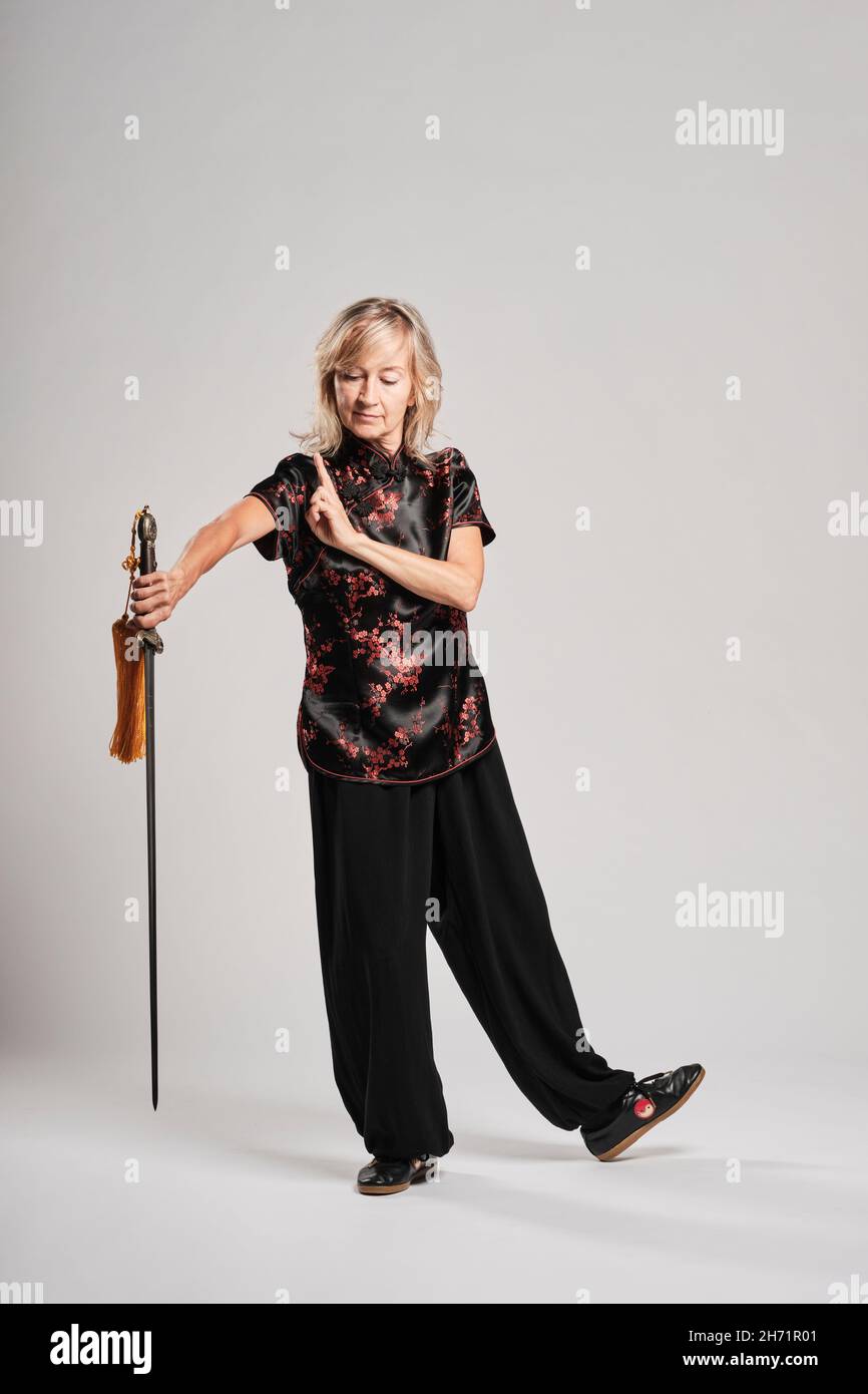 Reife blonde Frau, die Tai Chin mit Schwert auf weißem Hintergrund praktiziert, trägt eine traditionelle chinesische schwarze Jacke mit roter chinesischer Dekoration, schwarz Stockfoto