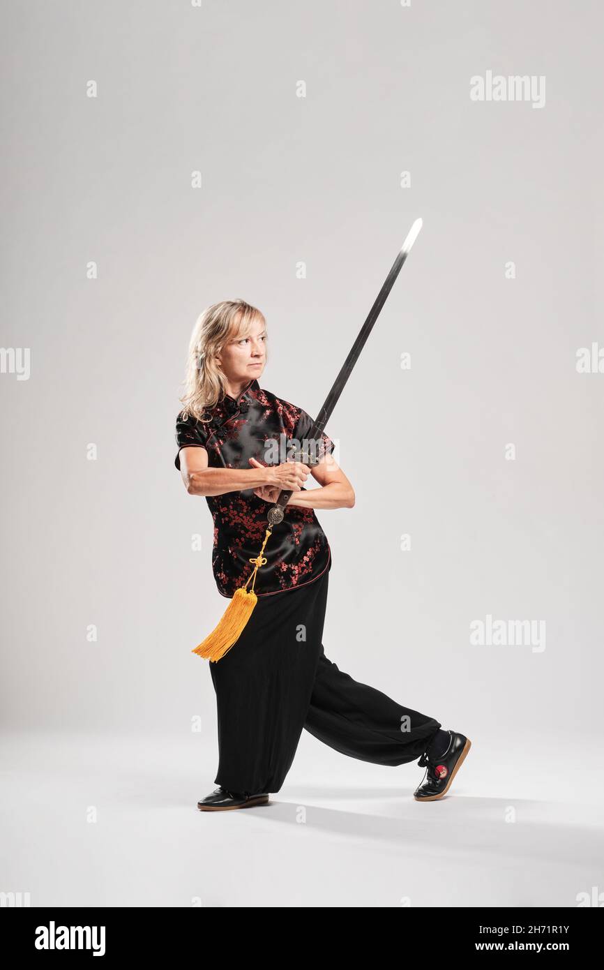 Reife blonde Frau, die Tai Chin mit Schwert auf weißem Hintergrund praktiziert, trägt eine traditionelle chinesische schwarze Jacke mit roter chinesischer Dekoration, schwarz Stockfoto