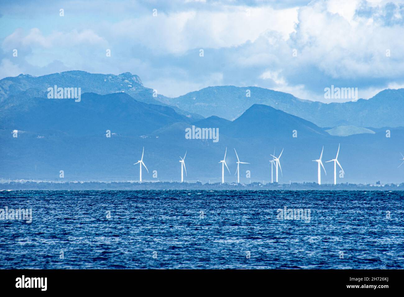 Dramatische Ansicht von weißen Windturbinen auf einem Hintergrund von blauen Bergen vom blauen Wellenwasser der Karibik aus gesehen. Puerto Rico, USA Stockfoto