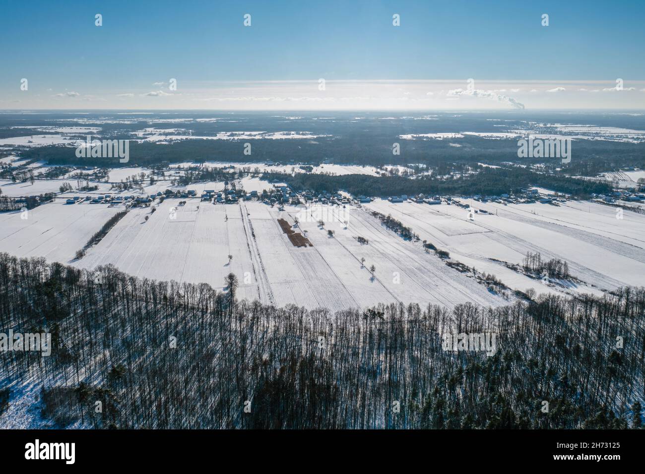 Winterlandschaft, Luftaufnahme des Waldes, schneebedeckte Felder und entferntes Dorf unter blauem Himmel Stockfoto