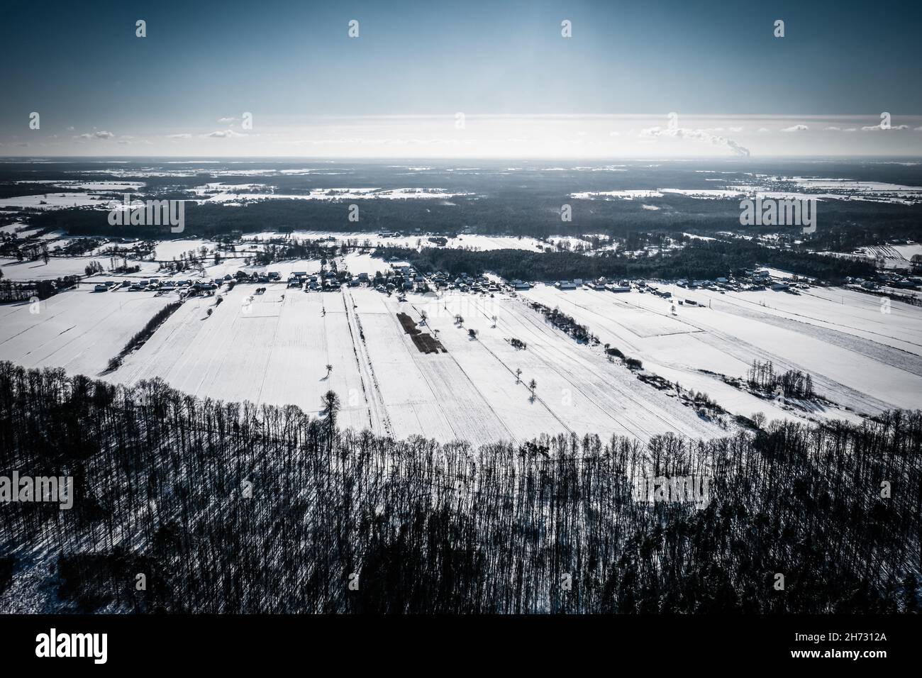 Winterlandschaft, Luftaufnahme des Waldes, schneebedeckte Felder und entferntes Dorf unter blauem Himmel Stockfoto