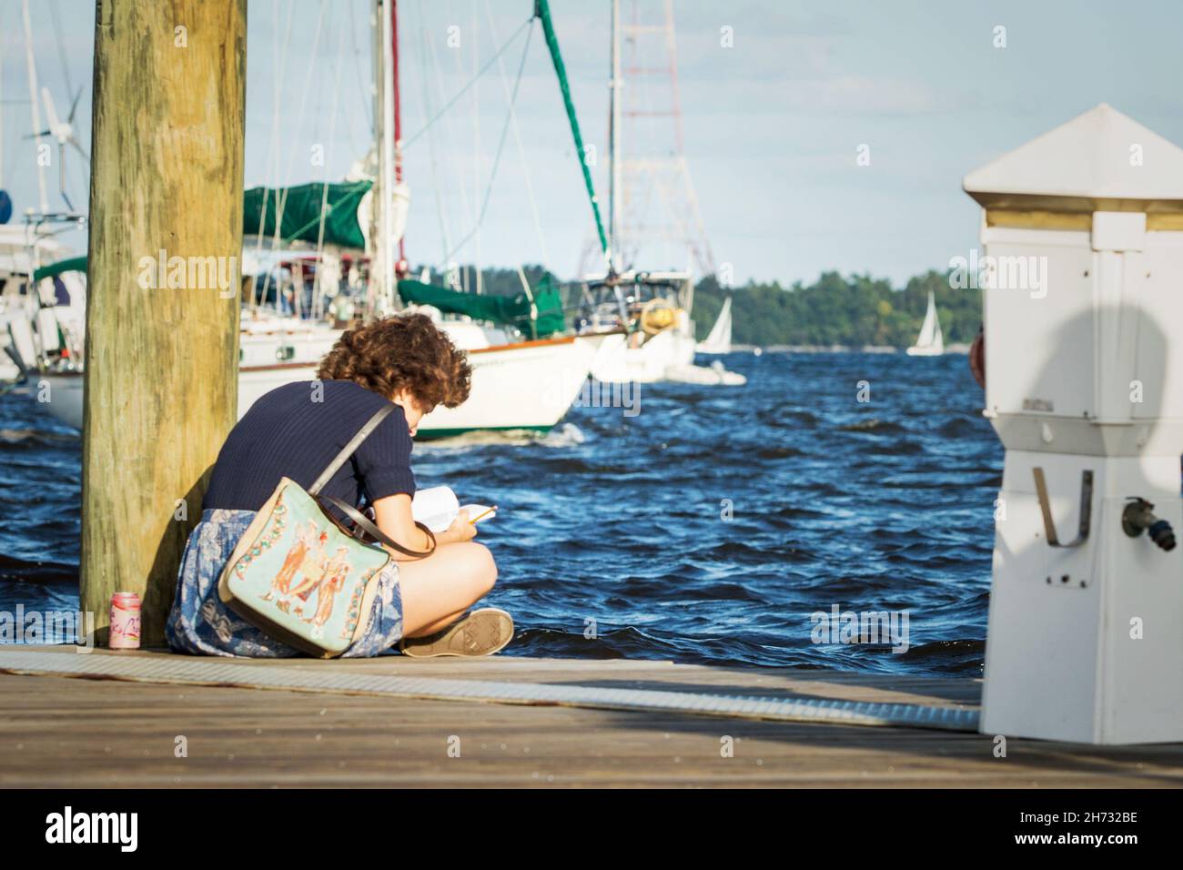 Eine junge Dame liest friedlich, während sie auf dem Dock der Ego Alley in der Annapolis Maryland Marina sitzt. Die Stimmung ist ruhig, während Segelboote an ihr vorbeifahren. Stockfoto