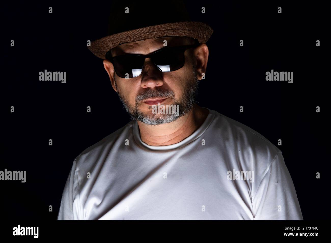 Bärtiger Mann Modell mit Hut und Sonnenbrille posiert ernst vor dunklem Hintergrund. Salvador, Bahia, Brasilien. Stockfoto
