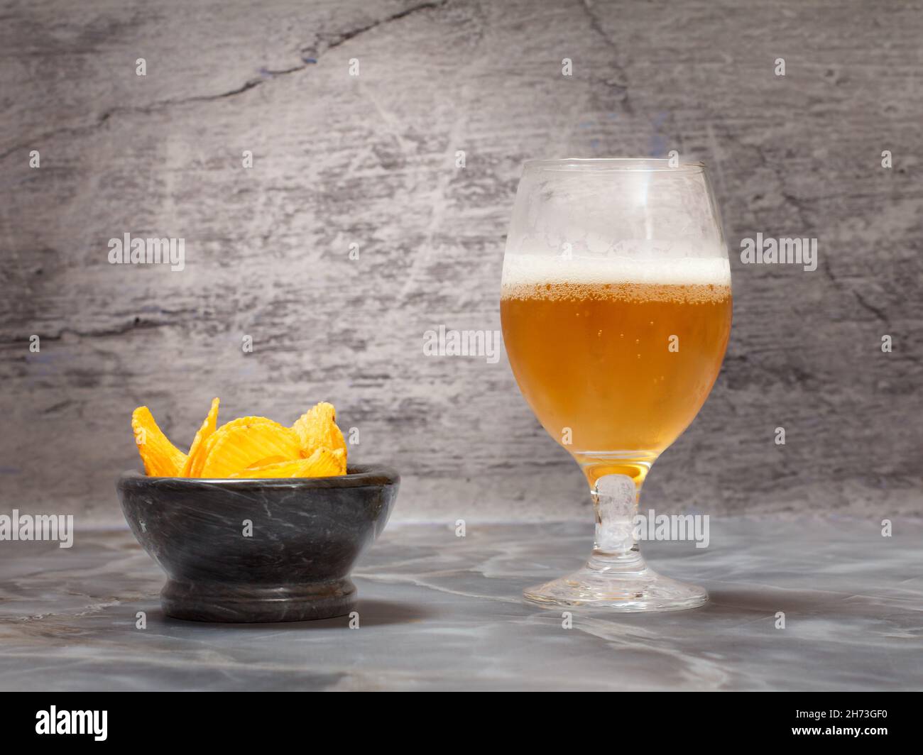 Ein Glas Bier und eine Steinschale mit Kartoffelchips auf grauem Hintergrund. Stockfoto