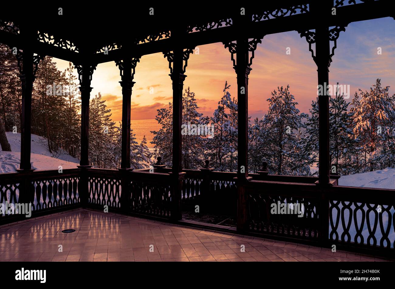 Blick auf den Sonnenuntergang über gefrorenem verschneiten Wald mit schneebedeckten Tannenbäumen vom Holzbalkon. Hölzerne durchbrochene Galerie oder Pavillon. Stockfoto
