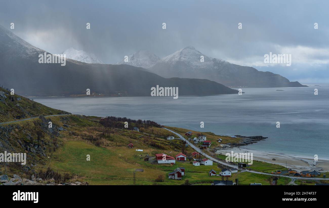 Bucht von Grøtfjord mit der Insel Vengsøya im Hintergrund. Herbst in Troms, Norwegen, gelbe Wiesen, verschneite Berge und bunte Häuser. Sturmfront Stockfoto