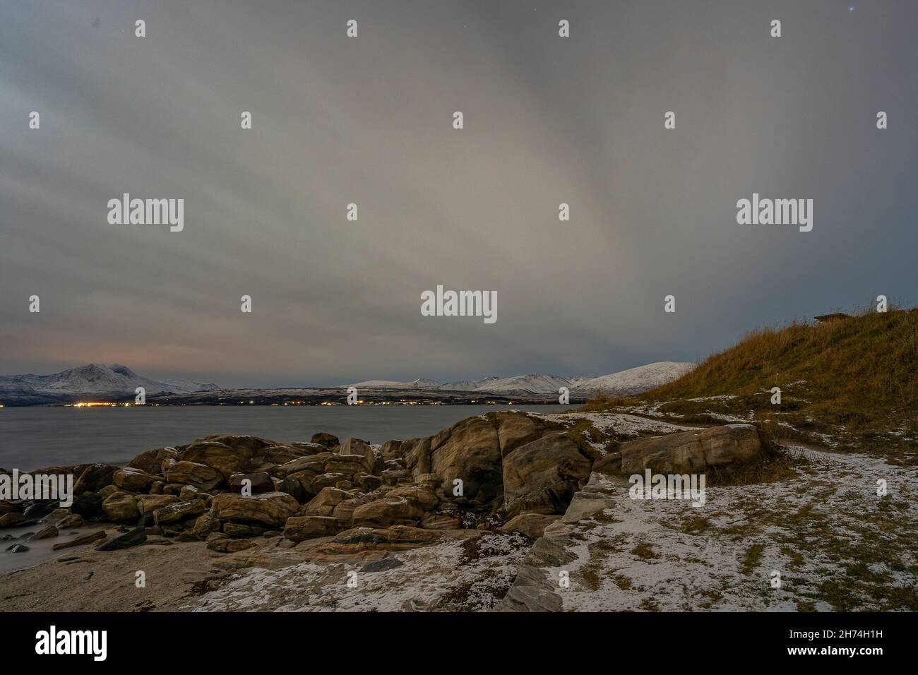 Telegraphbukta, Tromsø, Norwegen, mit wolkigem Himmel bei Nacht. Bucht mit Felsen, Kies und Gras, Rettungsring und erster Schnee im Herbst. Fjord Meer Stockfoto