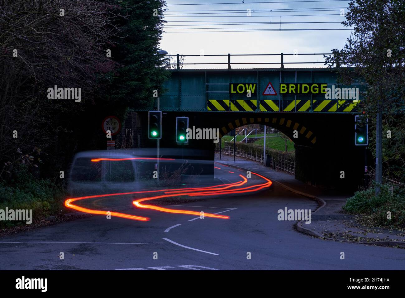 Autos mit Rotlichtwegen unter Eisenbahnbrücke mit niedrigem Brückenwarnschild in der Dämmerung Stockfoto