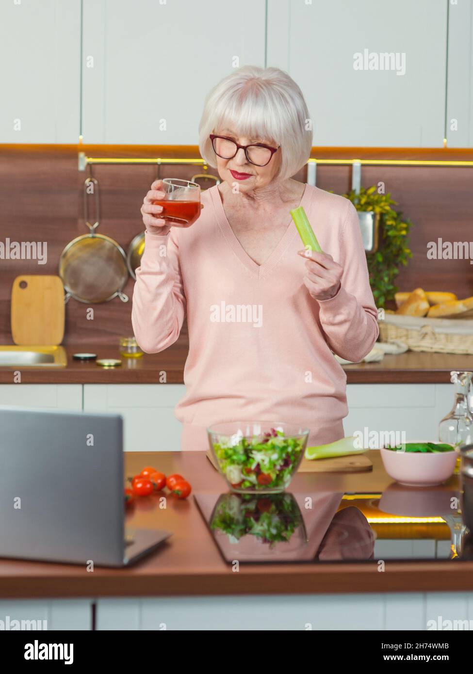 Ältere, fröhliche Videobloggerin, die Sellerie und Tomatensaft in ihrer Küche zeigt. Roh, vegetarisch, Diät, Blogger, Ernährungsberater Küche Konzept Stockfoto