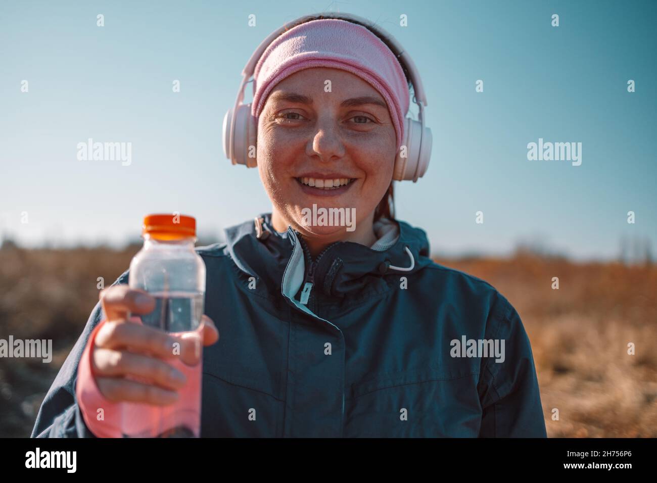 Aktive junge lächelnde Mädchen hält saubere Wasserflasche in der Hand, während sie in der Natur nach dem Training an einem sonnigen Tag in einem Herbstpark steht. Gesunder Lebensstil Stockfoto